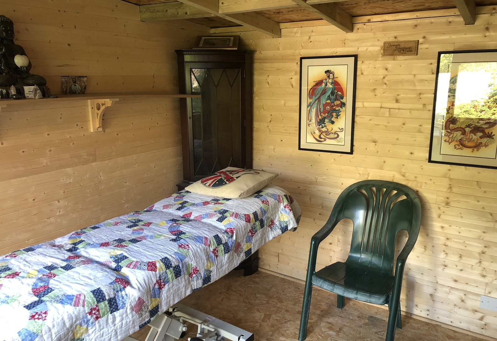 Inside the timber healing studio in Downend: a treatment couch with a patchwork quilt, a seated figure of the Buddha on a shelf and framed prints on pine walls.